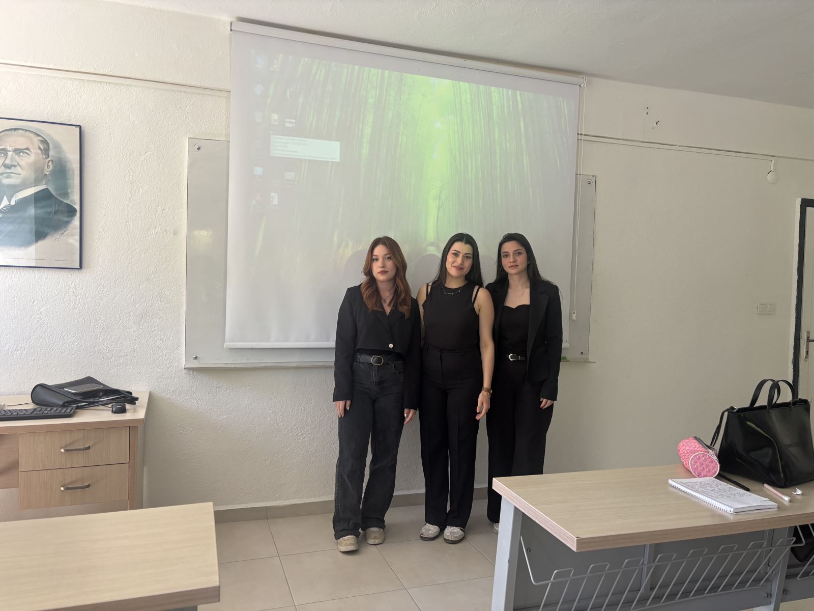 Bir sınıf ortamında üç kadın öğrenci projeksiyon perdesinin önünde yan yana durarak fotoğraf çekilmektedir. Öğrenciler koyu renk kıyafetler giymiştir ve kameraya doğru bakmaktadır. Arkada projeksiyon perdesine bilgisayar ekranı yansıtılmıştır. Sınıfta sıralar, öğretmen masası ve duvarda Atatürk portresi görülmektedir. Fotoğraf, öğrencilerin sunum etkinliği sonrasında çekilmiş bir hatıra karesi izlenimi vermektedir.